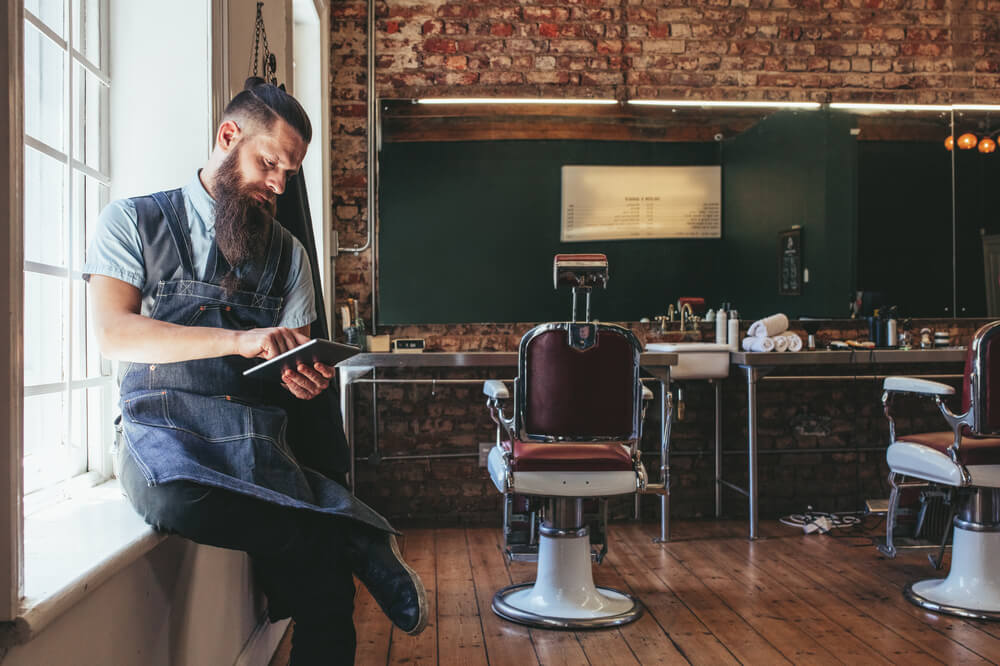 Barber With Digital Tablet Sitting On Window Sill Of Salon.